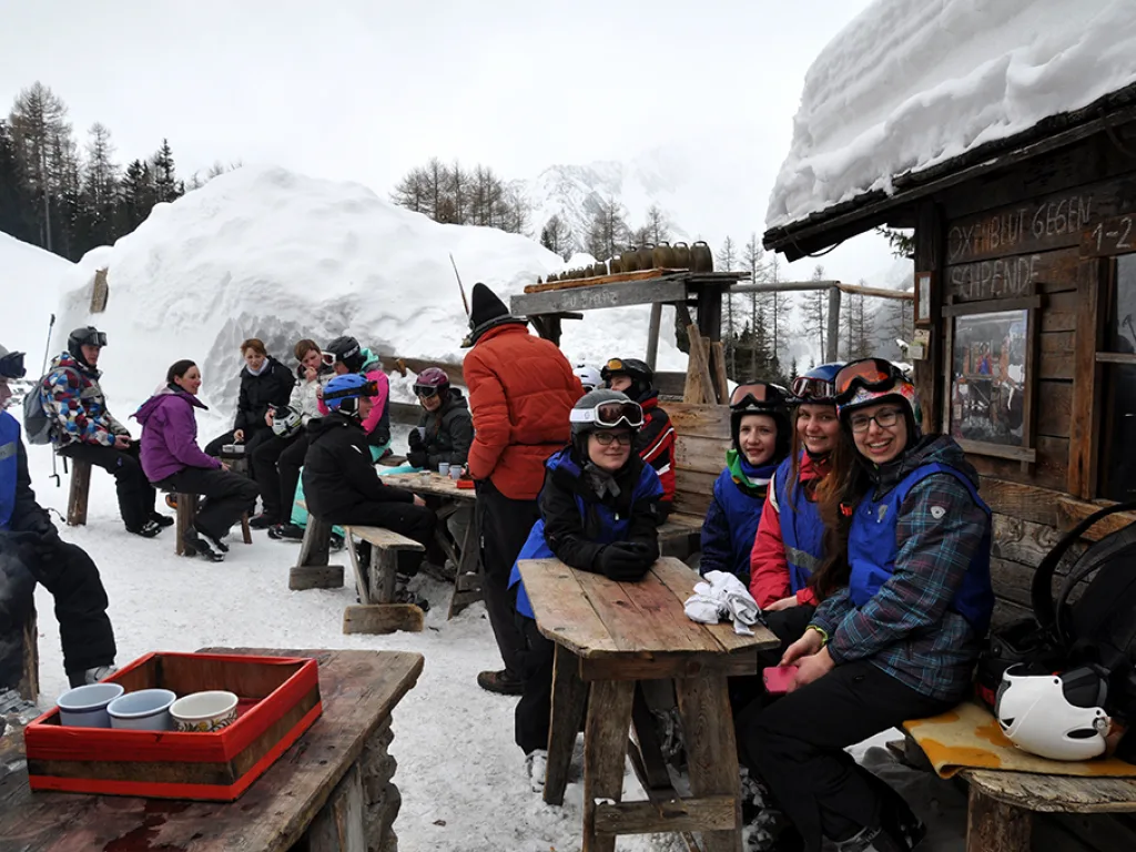 Jugendliche sitzen an Tischen vor einer eingeschneiten Hütte.